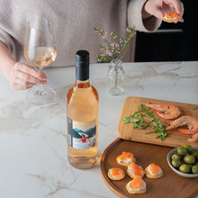 Load image into Gallery viewer, Bottle of rose wine on a marble countertop with a person holding a glass, a cutting board with shrimp and greens, and a small bowl of olives.