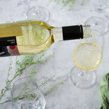 Load image into Gallery viewer, White wine being poured into a glass with a bottle and additional glasses on a light surface.