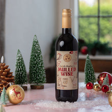 Load image into Gallery viewer, A bottle of Jacob Marley's Mulled Wine stands on a pink tabletop decorated with miniature Christmas trees, ornaments, and artificial snow, with a softly blurred indoor window background.