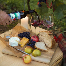 Load image into Gallery viewer, Person pouring wine into a glass with a charcuterie board featuring bread, cheese, and fruits under grapevines.