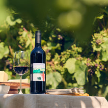Load image into Gallery viewer, Wine bottle and glass on a table with greenery in the background