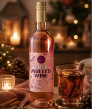 Load image into Gallery viewer, A bottle of Rose Mulled Wine stands on a wooden table beside a glass mug filled with a warm drink and cinnamon stick, surrounded by holiday lights, pinecones, and berries in a cozy festive setting.