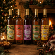 Load image into Gallery viewer, Five bottles of mulled cider arranged on a wooden table in a warm, festive setting with a fireplace, Christmas tree, and pinecones. The labels feature colorful illustrations of seasonal fruits including grapes, oranges, and pears.