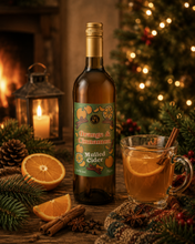 Load image into Gallery viewer, A bottle of Orange & Cinnamon Mulled Cider is displayed on a rustic wooden table surrounded by festive holiday decorations, including a lit mug with an orange slice and cinnamon stick, an orange half, star anise, pine branches, a lantern, and a Christmas tree in the background.