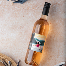 Load image into Gallery viewer, Bottle of rose wine with a scenic label on a textured surface with a hat and magazine.
