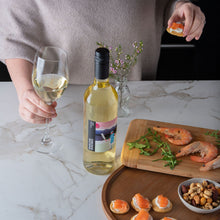 Load image into Gallery viewer, Person holding a glass of white wine with a bottle and food on a marble countertop
