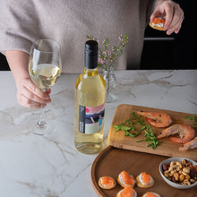 Load image into Gallery viewer, Person holding a glass of white wine with a bottle and food on a marble countertop
