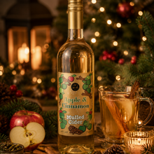 Load image into Gallery viewer, A bottle of Apple &amp; Cinnamon Mulled Cider by Celtc Marches is displayed on a rustic wooden table surrounded by festive holiday decorations, including a glass mug filled with the warm golden drink, cinnamon sticks, fresh apples, pinecones, and a glowing candle, with a blurred Christmas tree in the background.
