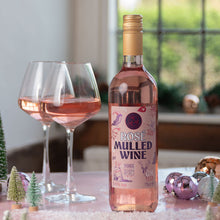 Load image into Gallery viewer, A bottle of Rose Mulled Wine stands on a decorated table with two filled wine glasses, small Christmas trees, and shiny ornaments in a softly lit indoor setting.
