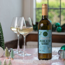 Load image into Gallery viewer, A bottle of White Mulled Wine stands on a pink tabletop with two filled wine glasses, small decorative trees, and a softly blurred indoor window background.
