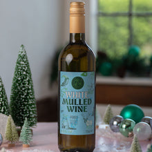 Load image into Gallery viewer, A bottle of White Mulled Wine stands on a pink surface with decorative miniature trees, snow-like flakes, and holiday ornaments in a softly lit indoor setting.
