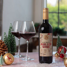 Load image into Gallery viewer, A bottle of Jacob Marley Mulled Wine stands on a tabletop beside two filled wine glasses, with small Christmas trees, ornaments, and a softly blurred window in the background.
