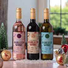 Load image into Gallery viewer, Three bottles of mulled wine arranged on a tabletop with holiday decorations. The left bottle is rosé wine with a pink label, the center bottle is dark red wine with a beige label, and the right bottle is white wine with a light blue label.
