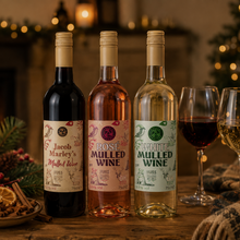 Load image into Gallery viewer, Three bottles of mulled wine in different colors are lined up on a wooden table with two wine glasses, candles, pinecones, and a cozy holiday fireplace background.
