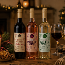 Load image into Gallery viewer, Three bottles of mulled wine in different colors are lined up on a wooden table with two wine glasses, candles, pinecones, and a cozy holiday fireplace background.

