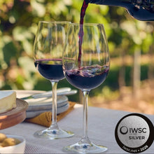 Load image into Gallery viewer, Red wine being poured into two glasses with a blurred vineyard background and IWSC Silver label.
