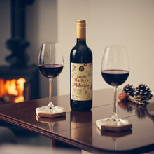 Load image into Gallery viewer, A bottle of mulled wine with two filled wine glasses on a dark wooden table in a cozy indoor setting, with a warm fireplace blurred in the background.
