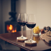 Load image into Gallery viewer, Two stemmed wine glasses filled with Radlow Hundred red wine on a wooden table, set in a cozy indoor scene with a blurred fireplace and warm ambient lighting.
