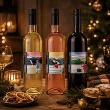 Load image into Gallery viewer, Three wine bottles with illustrated holiday labels are displayed on a rustic wooden table, surrounded by wine glasses, a candle, dried orange slices, and pastries, with a warm, festive Christmas tree in the background.
