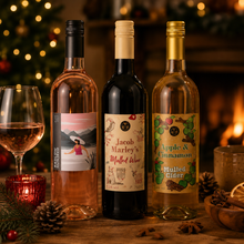 Load image into Gallery viewer, Three beverage bottles are arranged on a wooden table with a warm holiday fireplace and blurred lights in the background. A glass of rosé wine, mulled wine and apple and cinnamon mulled cider.
