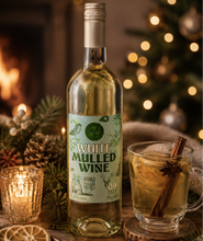 Load image into Gallery viewer, A bottle of White Mulled Wine stands on a rustic wooden surface beside a glass mug filled with a warm spiced drink and cinnamon stick, with candles, pinecones, and a softly blurred Christmas tree in the background.
