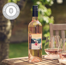 Load image into Gallery viewer, Bottle of rose wine with a scenic label on a wooden table outdoors, featuring a silver award badge.
