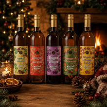 Load image into Gallery viewer, Five bottles of holiday-mixed white wine arranged on a wooden table in a warm, festive setting with a fireplace, Christmas tree, and pinecones. The labels feature colorful illustrations of seasonal fruits including grapes, oranges, and pears.
