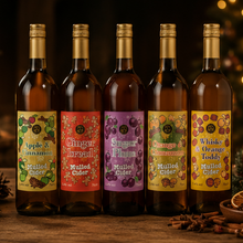 Load image into Gallery viewer, Five bottles of mulled cider arranged on a wooden surface with holiday decorations, including a lantern, pine branches, cinnamon sticks, dried orange slices, and a softly blurred Christmas tree with warm lights in the background.
